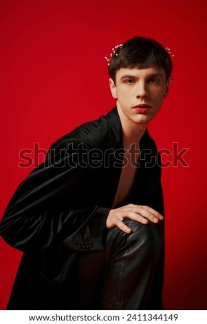 handsome man in black attire with leather pants and flowers in hair sitting on red background
