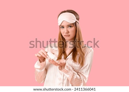 Upset young woman in pajamas taking pills on pink background