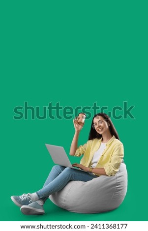 Young female programmer with laptop and cup of coffee sitting on grey beanbag chair against green background