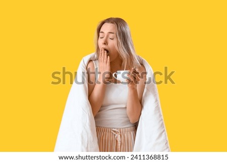 Sleepy young woman with blanket and cup of coffee on yellow background Royalty-Free Stock Photo #2411368815