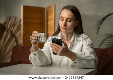A female customer uses a mobile phone, holding a box of medicines while sitting at a table.  Royalty-Free Stock Photo #2411389247