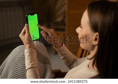 Close-up of a woman watching and using a smartphone on a green screen in the living room of the house. 