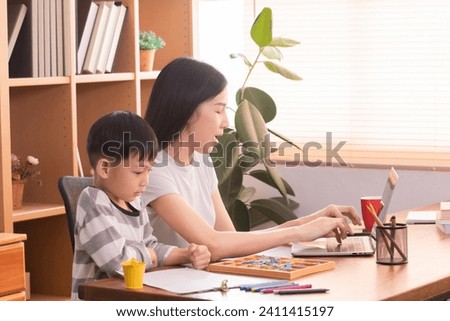 Happy Asian single mother multitask spent time playing alphabet toys and drawing with little boy while working freelance or creative design remote work from home, young teacher teaching preschool kid Royalty-Free Stock Photo #2411415197