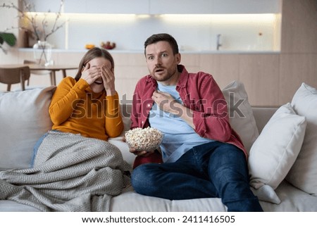 Couple spending time at home, watching TV, eating popcorn. Amazed intrigued husband looking with shoked interested glance. Embarrasing blushing wife closing eyes with hands of unexpected spicy episode Royalty-Free Stock Photo #2411416505
