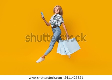 Full size photo of clever girl dressed print shirt jeans hold new outfit jumping hold smartphone isolated on yellow color background
