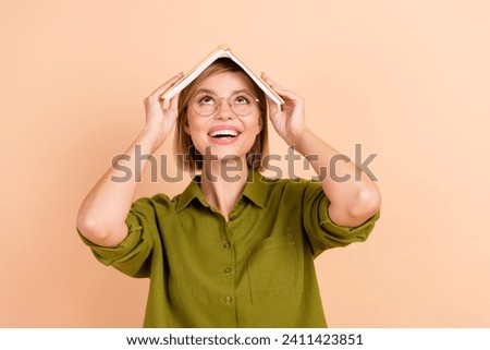 Photo of funny good mood woman dressed green shirt in glasses hold book on head look at offer empty space isolated on beige color background