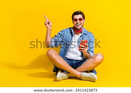 Full size photo of pleasant guy wear jeans jacket sit hold smartphone indicating at offer empty space isolated on yellow color background