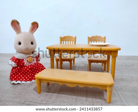 Adorable stuffed toy rabbit in red dress standing next to a miniature dining table on a white background, pretend play concept