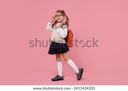 Happy schoolgirl in glasses with backpack on pink background Royalty-Free Stock Photo #2411434105