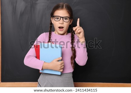 Surprised schoolgirl in glasses holding books near chalkboard
