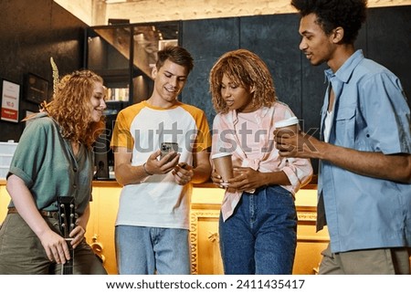 happy man showing smartphone to multicultural mates with coffee to go and guitar in lobby of hostel Royalty-Free Stock Photo #2411435417