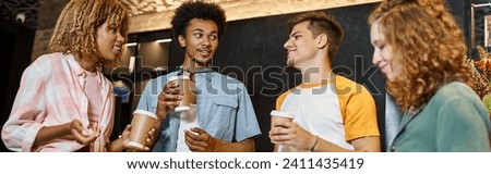 smiling and trendy multicultural buddies with paper cups talking in lobby of youth hostel, banner Royalty-Free Stock Photo #2411435419