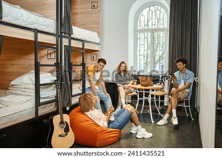 young and stylish multicultural buddies talking near laptop and acoustic guitar in cozy hostel room Royalty-Free Stock Photo #2411435521