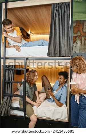 cheerful multicultural students talking on double-decker bed in cozy hostel room, travelers Royalty-Free Stock Photo #2411435581