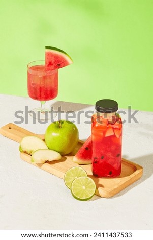 Green apples, lemons, fresh watermelon and a detox water bottle are displayed on a wooden tray on a white surface with a green background. Fresh fruit provides a rich amount of vitamins for the body.