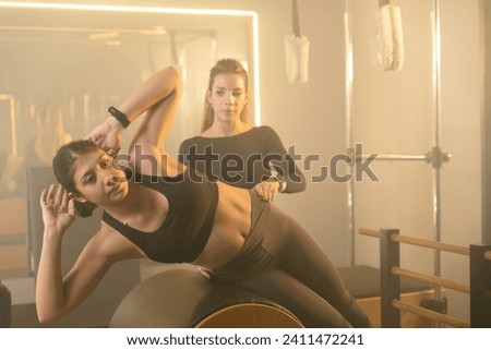 A young woman performs core exercises on a Reformer Pilates machine in a gym, guided by a trainer in the Reformer room