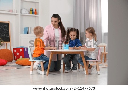 Nursery teacher and group of cute little children drawing at desk in kindergarten. Playtime activities Royalty-Free Stock Photo #2411477673