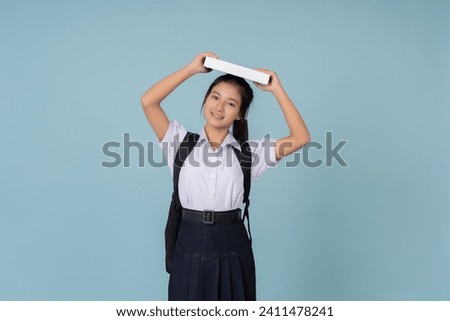 Asian female high school student Holding up a textbook and carrying a bag Present on the website or poster of the educational tutoring institute. Taking photos in a studio with a blue background