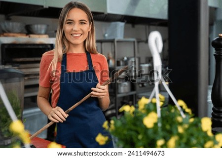 Smiling confident young woman chef looking at camera holding large wooden mixing spoon standing in commercial kitchen in restaurant. Young woman baker with wooden spoon in bakery.