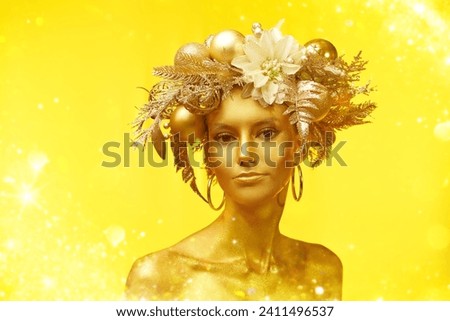 Christmas beauty portrait. Beautiful young woman with golden make-up and shiny golden skin poses in a Christmas wreath. Yellow studio background. Holiday luxury look. Copy space.