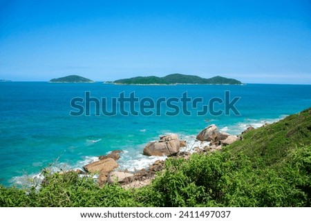 The picture shows a small island in the ocean and a rocky coastline with turquoise-green water, green slopes and an absolutely blue sky without clouds.