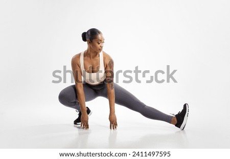 Fit black lady making extended leg squat pose stretching legs, exercising against white background. Full length studio shot of athlete woman having workout for muscles flexibility Royalty-Free Stock Photo #2411497595
