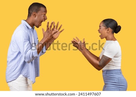 Black man and woman engage in playful gestures with exaggerated surprised expressions, creating humorous and lively scene on vivid yellow background Royalty-Free Stock Photo #2411497607