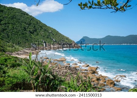 The picture shows a hidden, small sandy beach between rocks in a small bay on the mountainous slopes of an island covered in dense rainforest. Ilha de Santa Catarina, Brazil