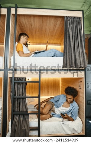 multicultural students using gadgets on double-decker beds in contemporary youth hostel, travelers Royalty-Free Stock Photo #2411514939