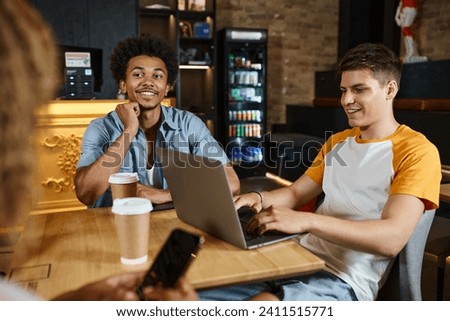 young smiling man networking on laptop near multicultural friends in lobby cafe of students hostel Royalty-Free Stock Photo #2411515771