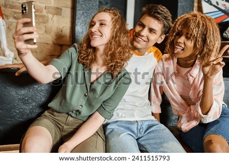 cheerful woman taking selfie with multiethnic friends while having fun in lobby of youth hostel Royalty-Free Stock Photo #2411515793