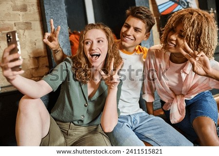 excited multicultural friends showing victory signs and taking selfie on smartphone in hostel lobby Royalty-Free Stock Photo #2411515821