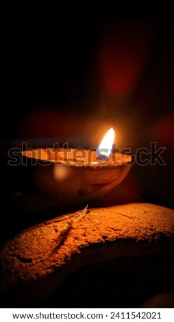 In the dark night, the clay oil lamp casts a soft, warm glow, creating an intimate ambiance. The flickering flame dances within, casting shadows that play on the surrounding surfaces. Royalty-Free Stock Photo #2411542021