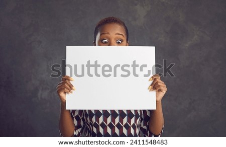 Funny surprised black woman showing empty white paper banner standing against grey background. Short haired young African lady peeking at clean blank mockup sign she's holding hiding half of her face