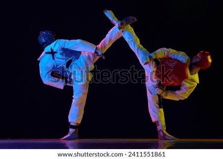 Two men in kimono and helmets practicing taekwondo, training, fighting against black background in neon light. Concept of martial arts, combat sport, competition, action, strength