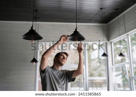 A man is captured in the act of changing a light bulb in a hanging ceiling light fixture, with natural daylight illuminating the scene. Royalty-Free Stock Photo #2411585865