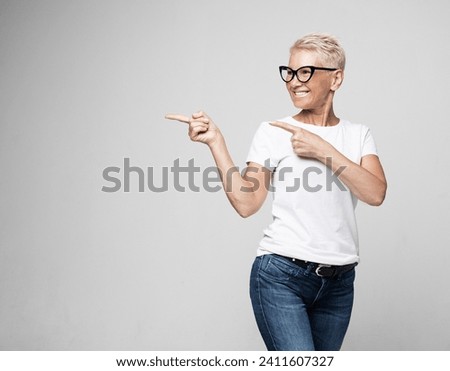 Smiling senior gray-haired woman isolated on grey studio background pointing with finger at blank copy space aside