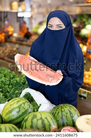 Young Muslim woman in traditional islamic dress and niqab covering face, holding quarter of ripe watermelon wrapped in film while standing among fruit display stands in store