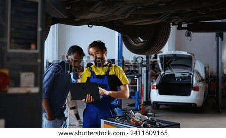 Trained mechanic and customer standing underneath car in garage using laptop to order new parts after finding defects. Employee assisting african american client by looking for components online Royalty-Free Stock Photo #2411609165