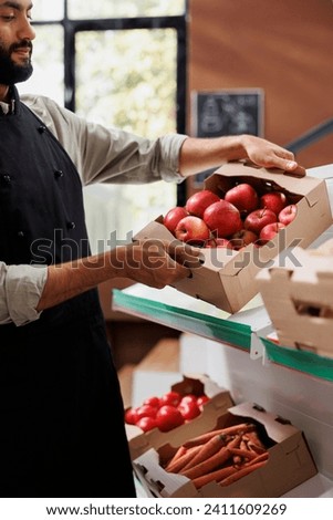 Close-up of merchant with a black apron picking up a crate of red apples from a shelf in environmentally conscious store. Male shopkeeper carrying box of locally grown fresh produce. Royalty-Free Stock Photo #2411609269