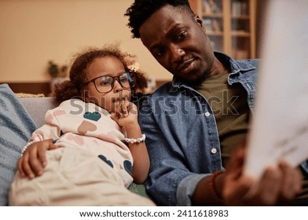 Medium shot of African American preschool girl in glasses on sofa at home interested in pictures of fairy tale book in fathers hands