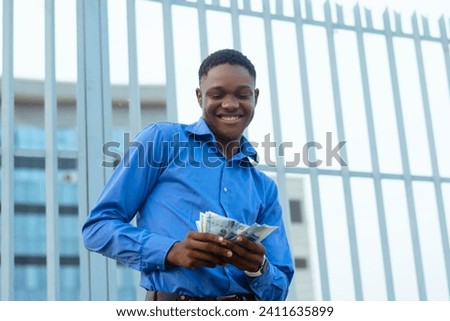 Excited handsome black man in a blue shirt is seen holding a sum of money in this stock photo illustrating a financial concept.