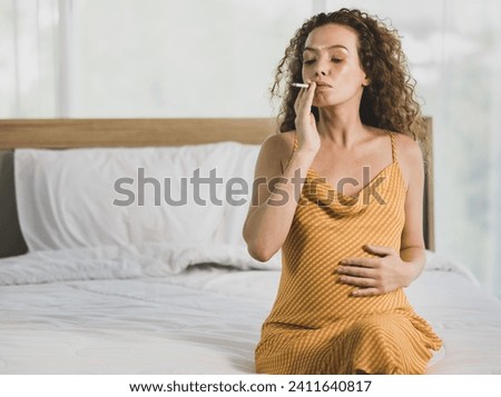 Curly hairstyle young unhappy unhealthy bad behavior Caucasian pregnancy mother in maternity long dress cloth sit on bed in bedroom holding smoking cigarette taking risk and danger to unborn child. Royalty-Free Stock Photo #2411640817