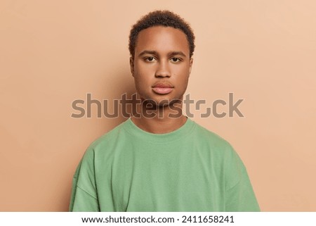 People emotions concept. Studio shot of young calm relaxed African american male with short curly hair wearing green casual tshirt standing in centre isolated on beige background keeping hands down