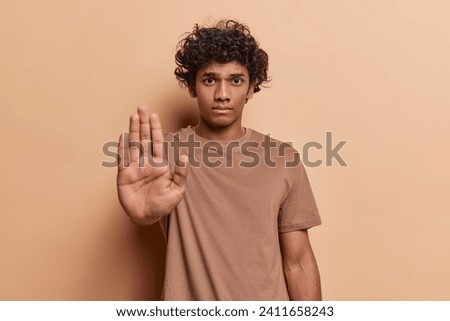 Waist up shot of curly haired Hindu man stretches out hand to block or refuse something shows stop gesture has serious strict expression dressed in casual t shirt isolated over brown background