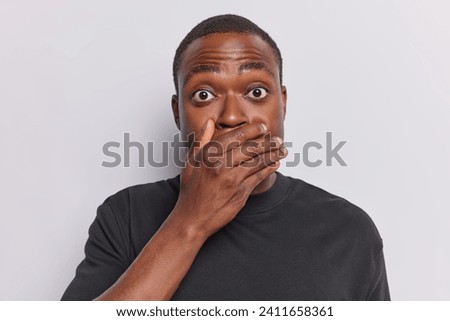 Secret concept. Dark skinned young man covers mouth with hand tries to be speechless has bugged eyes dressed in black t shirt isolated over white studio background standing intense and scared Royalty-Free Stock Photo #2411658361