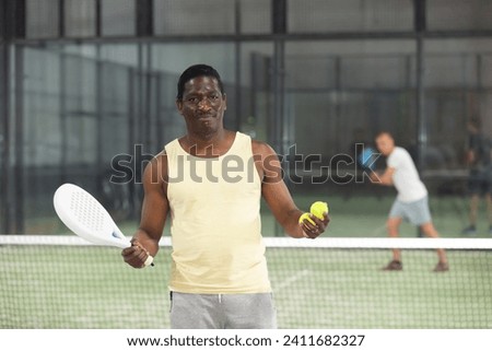 Portrait of positive afro american man with padel racket and tennis ball at court indoor