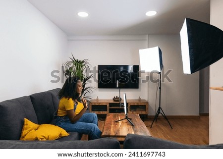 Side view of young black woman video blogger recording herself with smartphone, ring light and spotlights at home. She wears an overall and a yellow shirt. She smiles greeting her followers. 