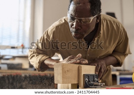 Black carpenter carefully brushing sawdust off wooden board while building furniture in workshop