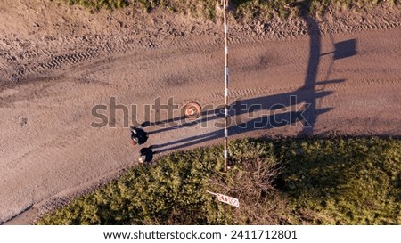 This aerial image captures a solitary figure at a crossroads, the long shadows of late afternoon stretching across the textured ground. The crossroads are marked by a traffic sign indicating a speed